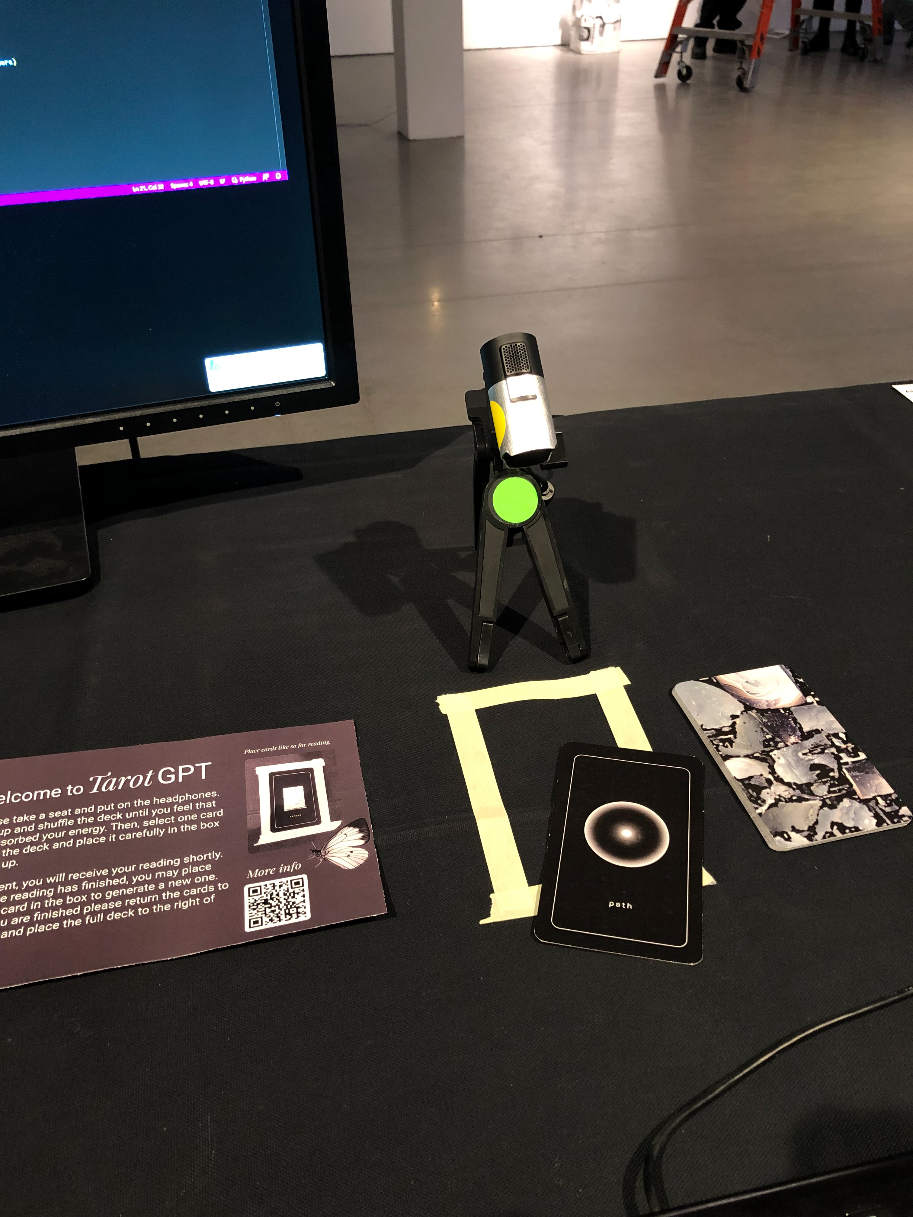 Detailed close up of a table with black table cloth, headphones, a keyboard, a mouse, playing cards and an instruction manual on top. There is a webcam connected to a 6 inch tripod pointing towards the cards.